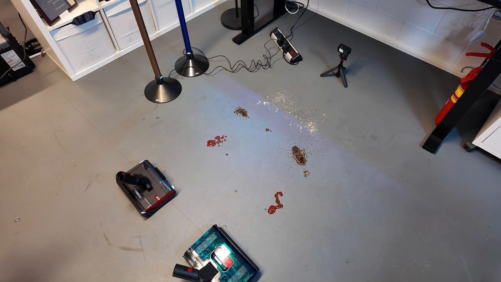 Grijze vloer met plekken ketchup, koffie en koekkruimels, met twee wasmonden en de dweilen in hun laadvoet