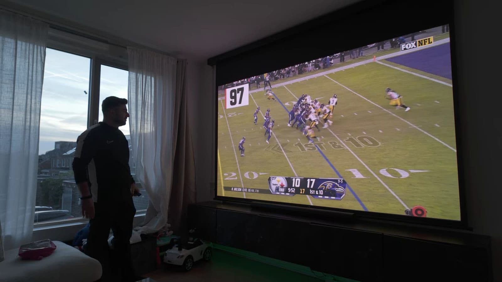 Full living room view during daytime with large windows and bright 120-inch projection showing an NFL game